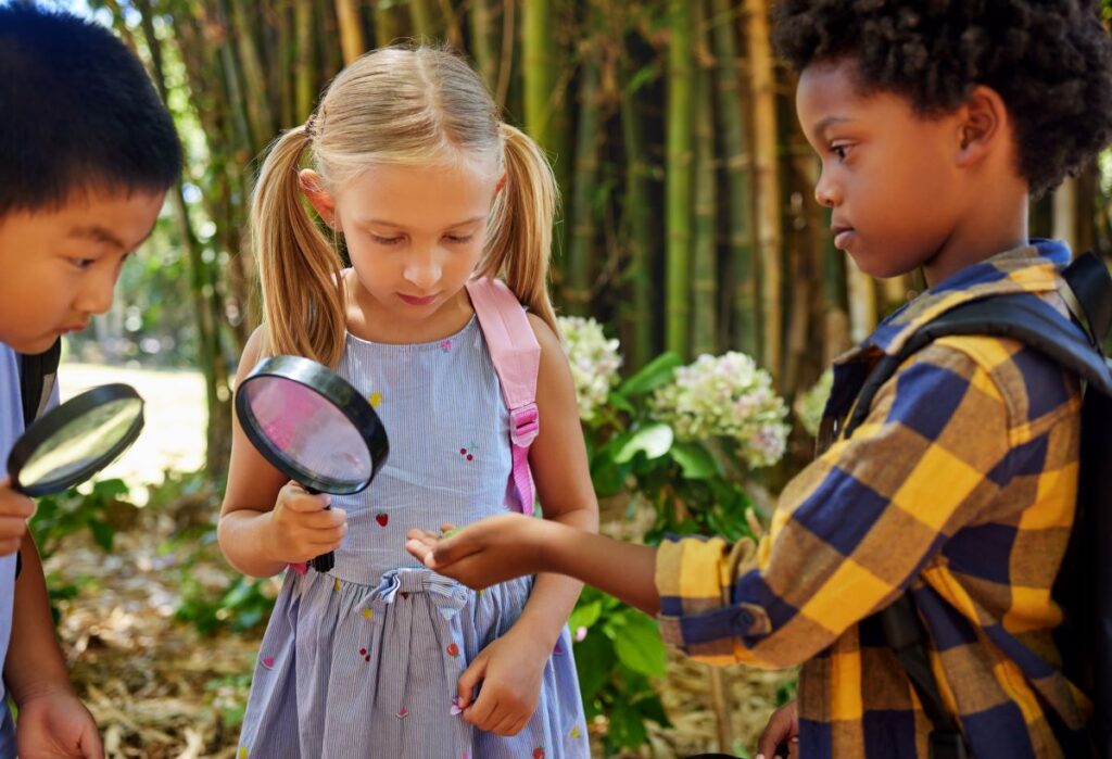 2 boys and 1 girl examine something in a boy's hand. 2 of the children are holding magnifying glasses.