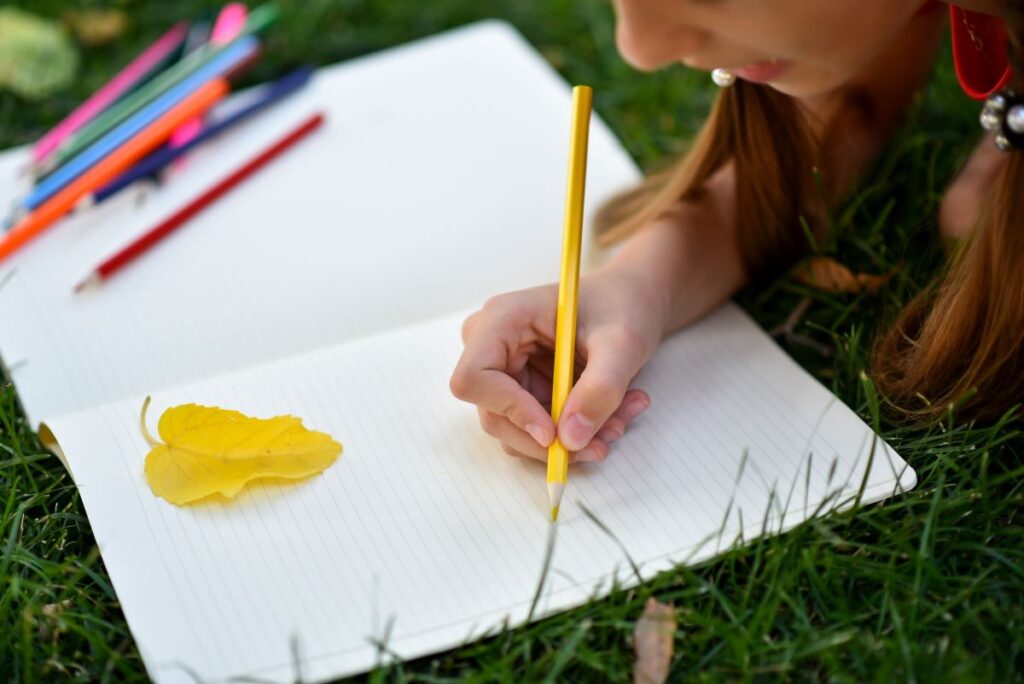 A girl is about to draw a yellow leaf sitting on her notebook. She is holding a yellow pencil.