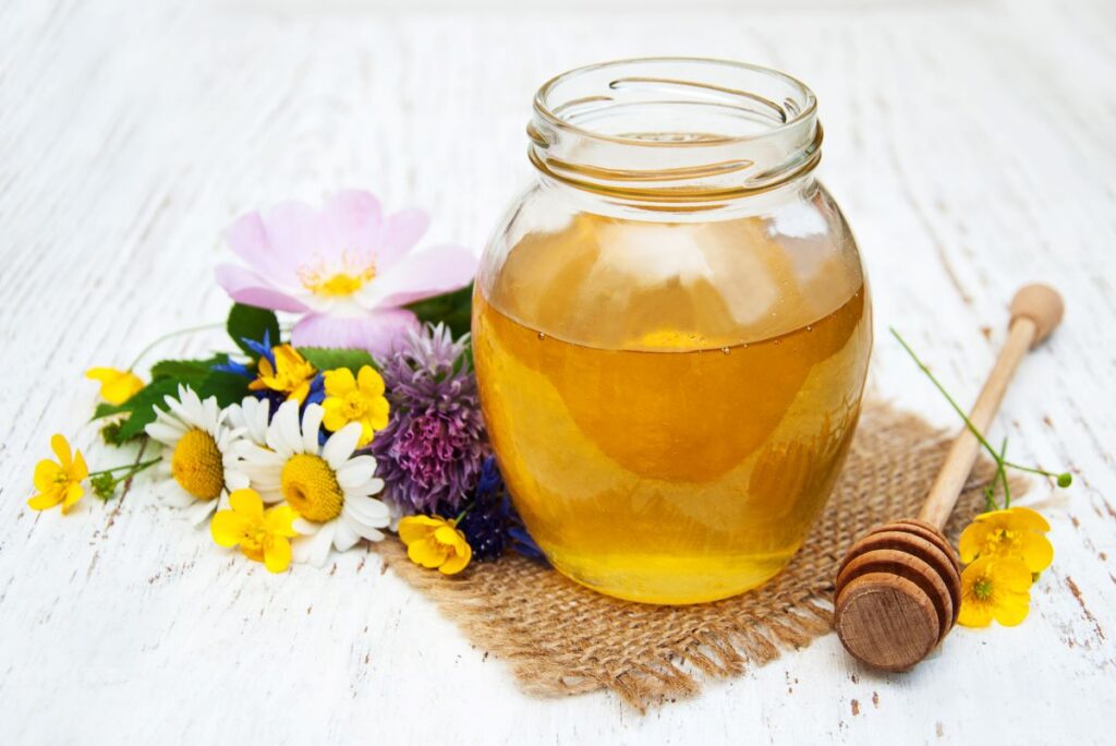 A jar of honey sitting on a burlap pad with wildflowers and a honey stirrer next to it.