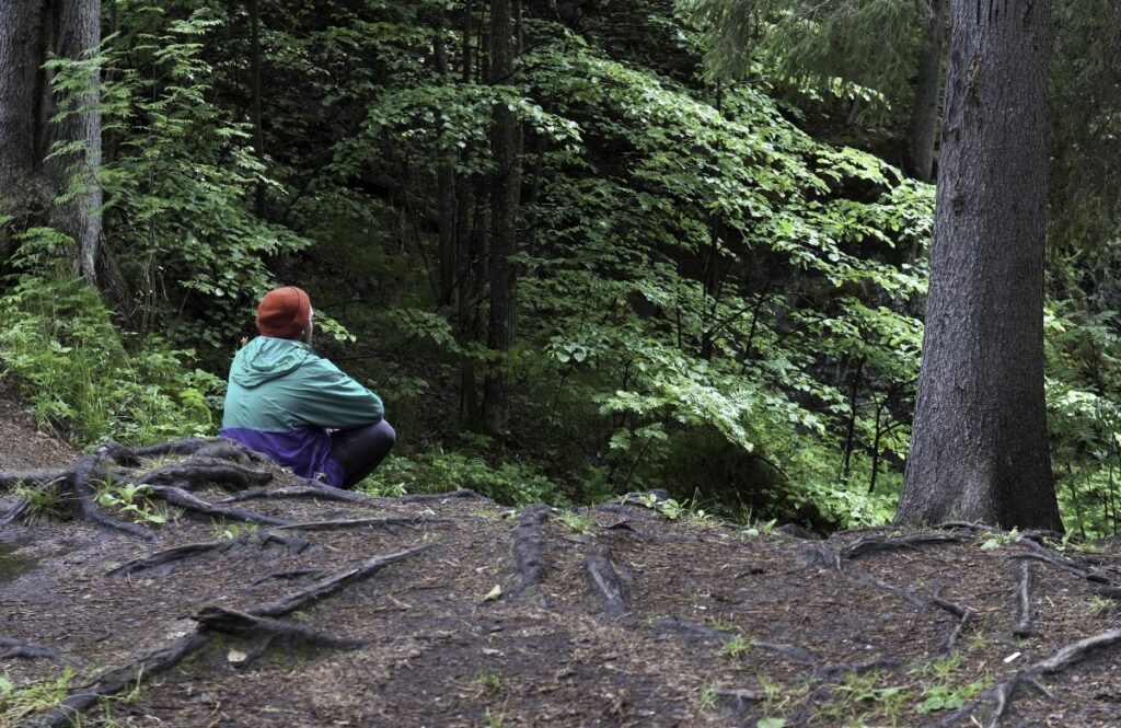 A man in a turquoise jacket and red hat sits on a trail in the woods.